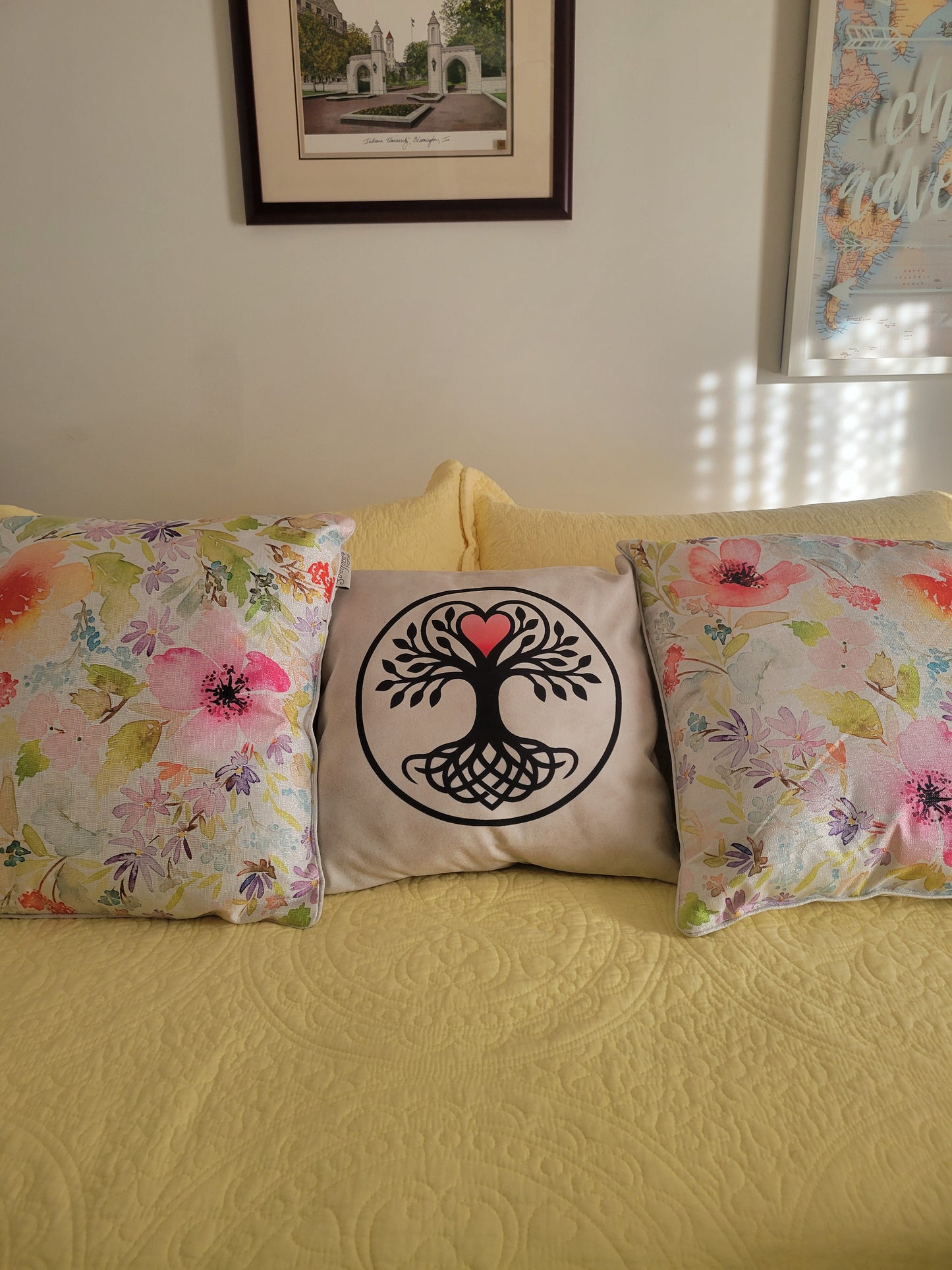 Black Tree of Life with a pink heart crown and knot-work roots in a black circle on a cream throw pillow, displayed on a yellow bedspread.