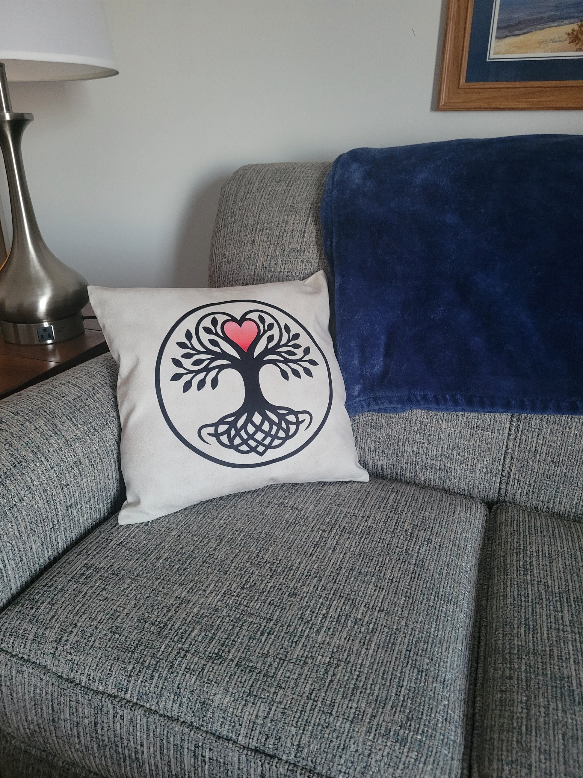 Cream throw pillow featuring a black Tree of Life with Celtic knot-work roots and a pink heart canopy, enclosed in a circle, displayed in the left corner of a gray sofa.