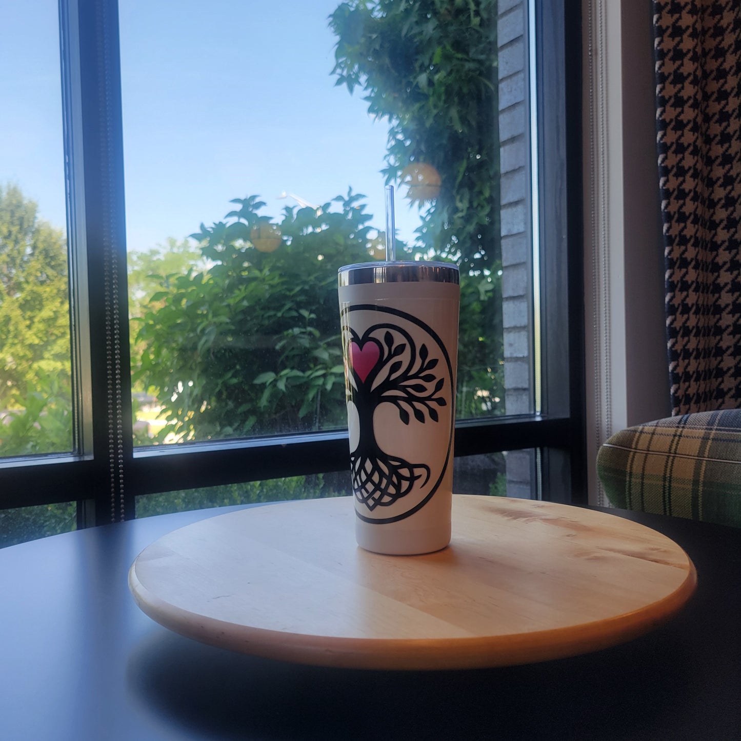 White stainless-steel tumbler with a black Norse-style Tree of Life graphic accented by a pink heart, displayed on a wooden tray by a sunlit window.