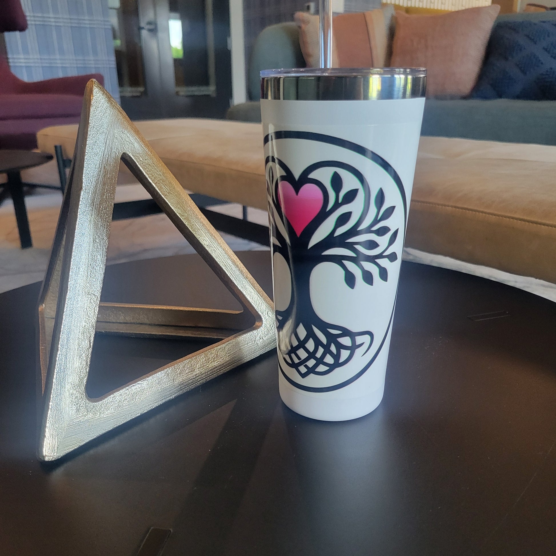 White stainless-steel tumbler featuring a black circular tree-of-life design with a pink heart at its center, displayed on a dark table beside a gold triangular decorative object.