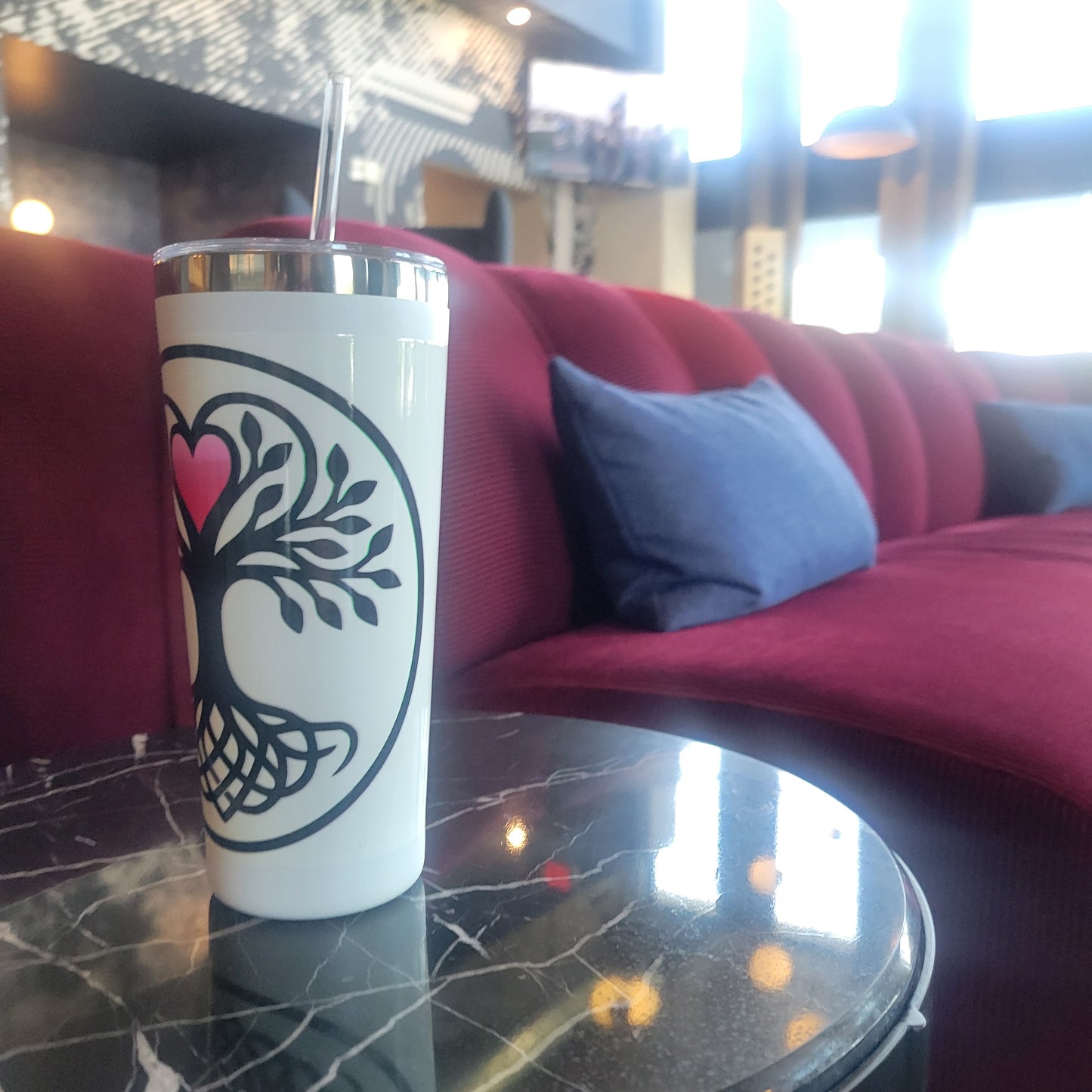 White tumbler with a Norse tree of life design featuring interwoven roots, black leafy branches, and a glowing pink heart, displayed on a marble table in front of a luxury sofa — drinkware.