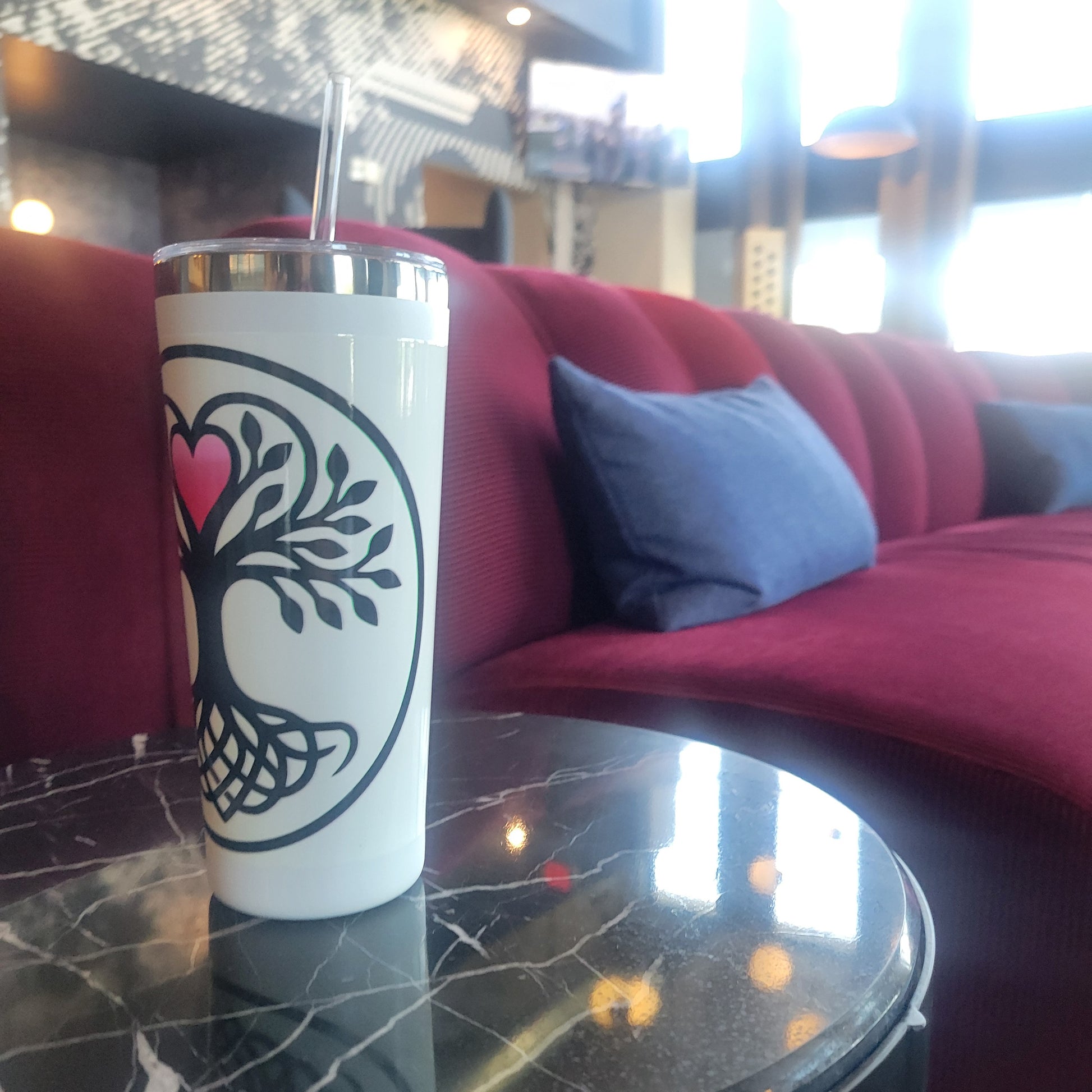 White tumbler with a Norse tree of life design featuring interwoven roots, black leafy branches, and a glowing pink heart, displayed on a marble table in front of a luxury sofa — drinkware.