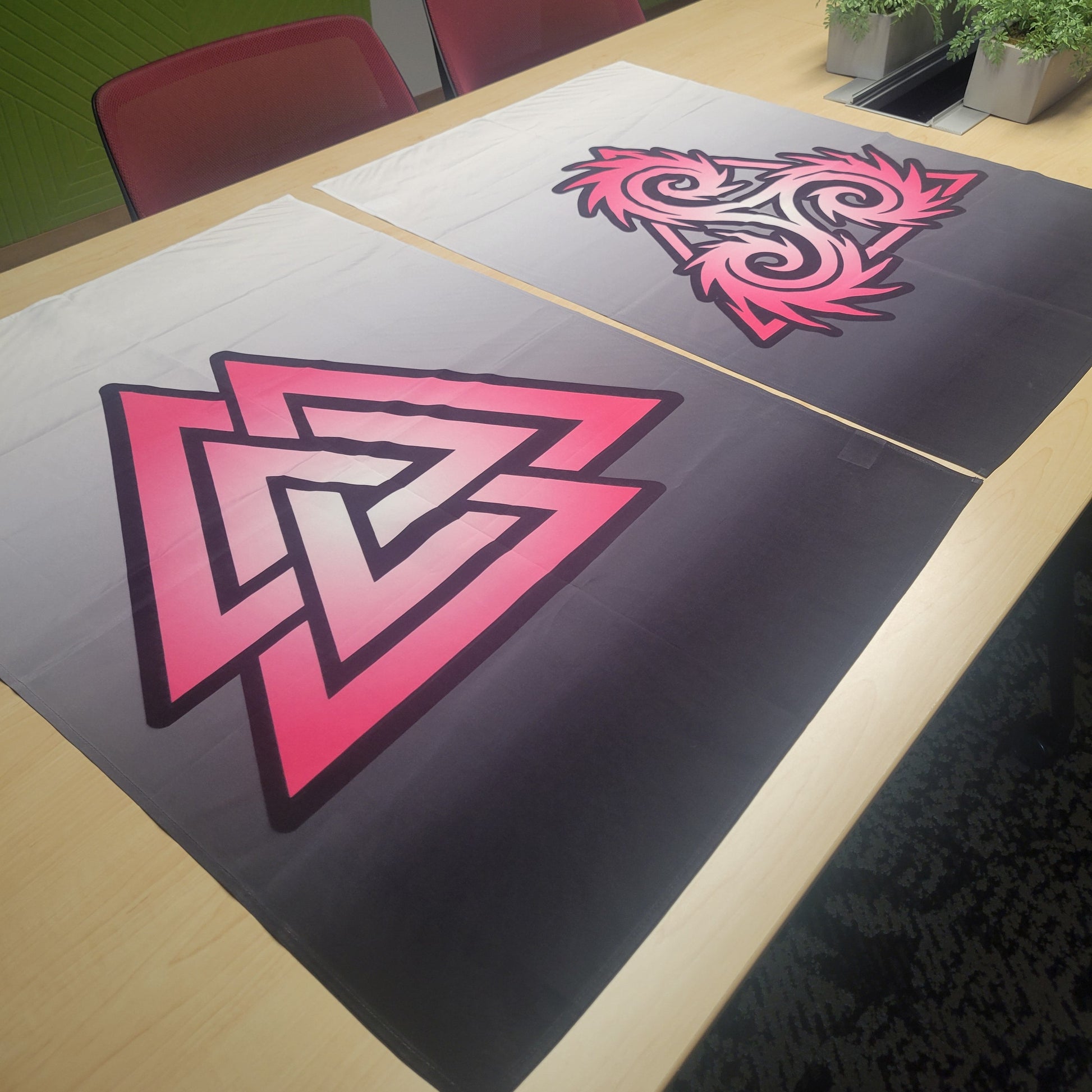 Two white-to-black tapestries laid flat on a table: one featuring a bold pink gradient Valknut symbol, the other a swirling pink triskelion design. Wall tapestries.
