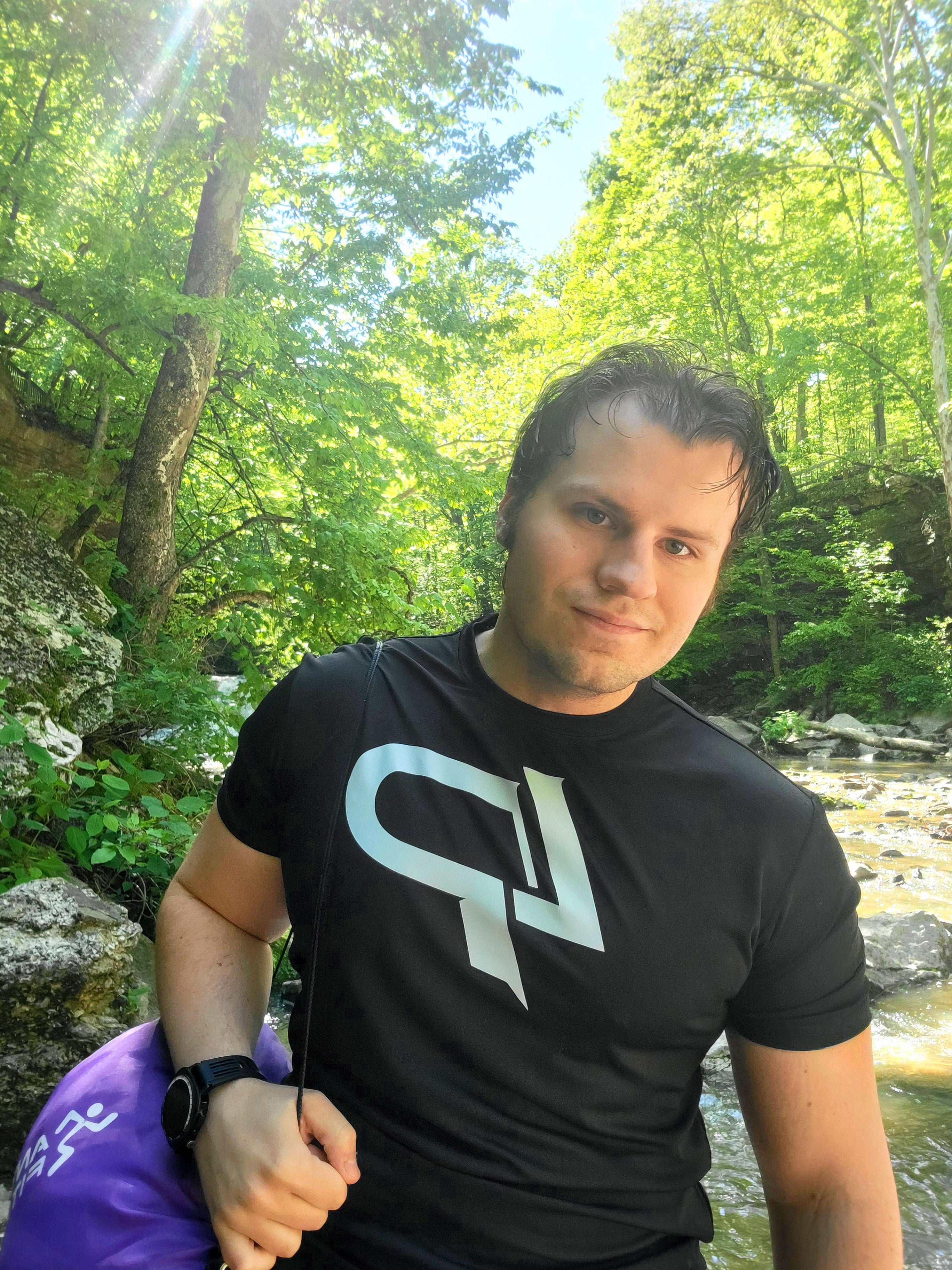 A photograph of Conner, the founder of SpiritForge smiling and wearing a black LP logo shirt while hiking in the woods next to a rushing river.